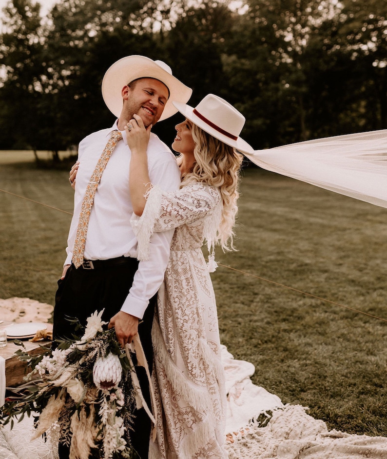 May include: A couple in a field, wearing white cowboy hats. The woman is wearing a white lace dress and a veil. The man is wearing a white shirt and black pants. They are holding a bouquet of flowers.