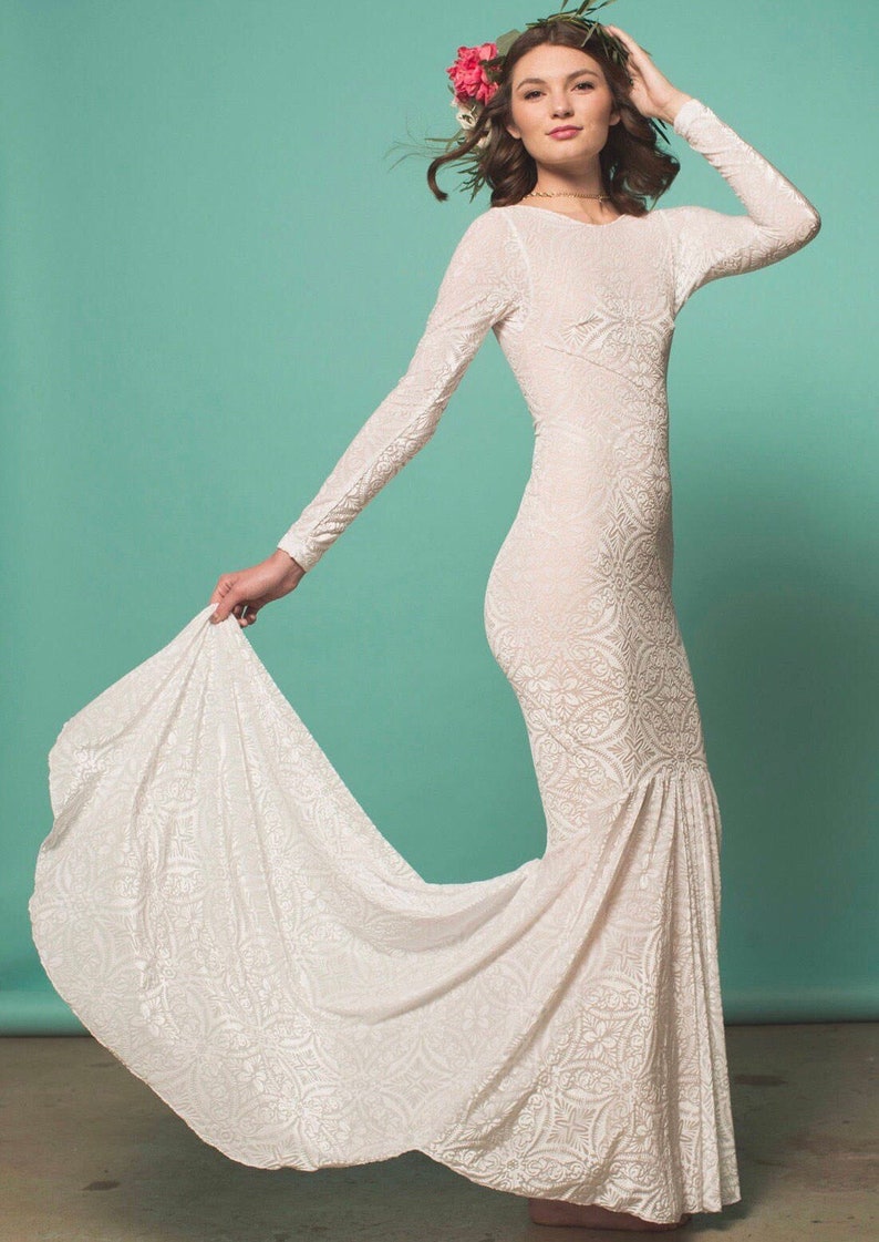 May include: A white, long-sleeved, lace wedding dress with a fitted bodice and a flowing skirt. The dress has a floral pattern and the model is wearing a flower crown.