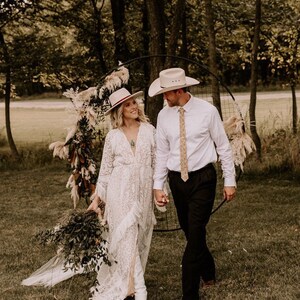 May include: A couple in a rustic outdoor setting, wearing white cowboy hats and wedding attire. The bride is wearing a white lace dress and the groom is wearing a white shirt and black pants. They are standing in front of a circular archway decorated with dried flowers and greenery.