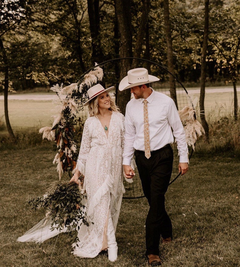 May include: A couple in a rustic outdoor setting, wearing white cowboy hats and wedding attire. The bride is wearing a white lace dress and the groom is wearing a white shirt and black pants. They are standing in front of a circular archway decorated with dried flowers and greenery.