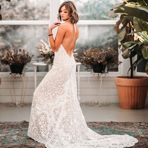May include: A white lace wedding dress with a halter neckline and a low back. The dress has a fitted bodice and a flowing skirt.