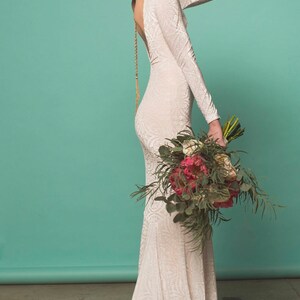 May include: A woman wearing a white lace wedding dress with a low back and long sleeves. She is wearing a flower crown and holding a bouquet of pink and white flowers.