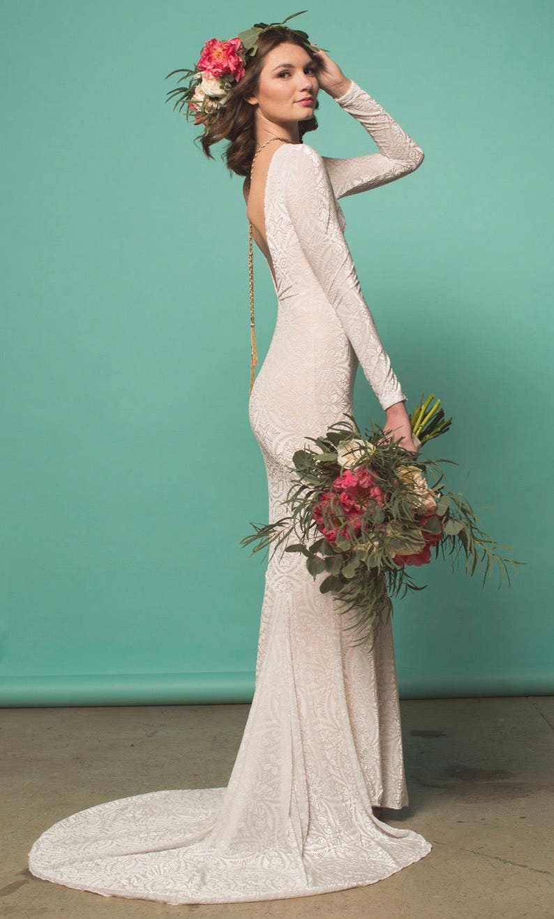 May include: A woman wearing a white lace wedding dress with a low back and long sleeves. She is wearing a flower crown and holding a bouquet of pink and white flowers.