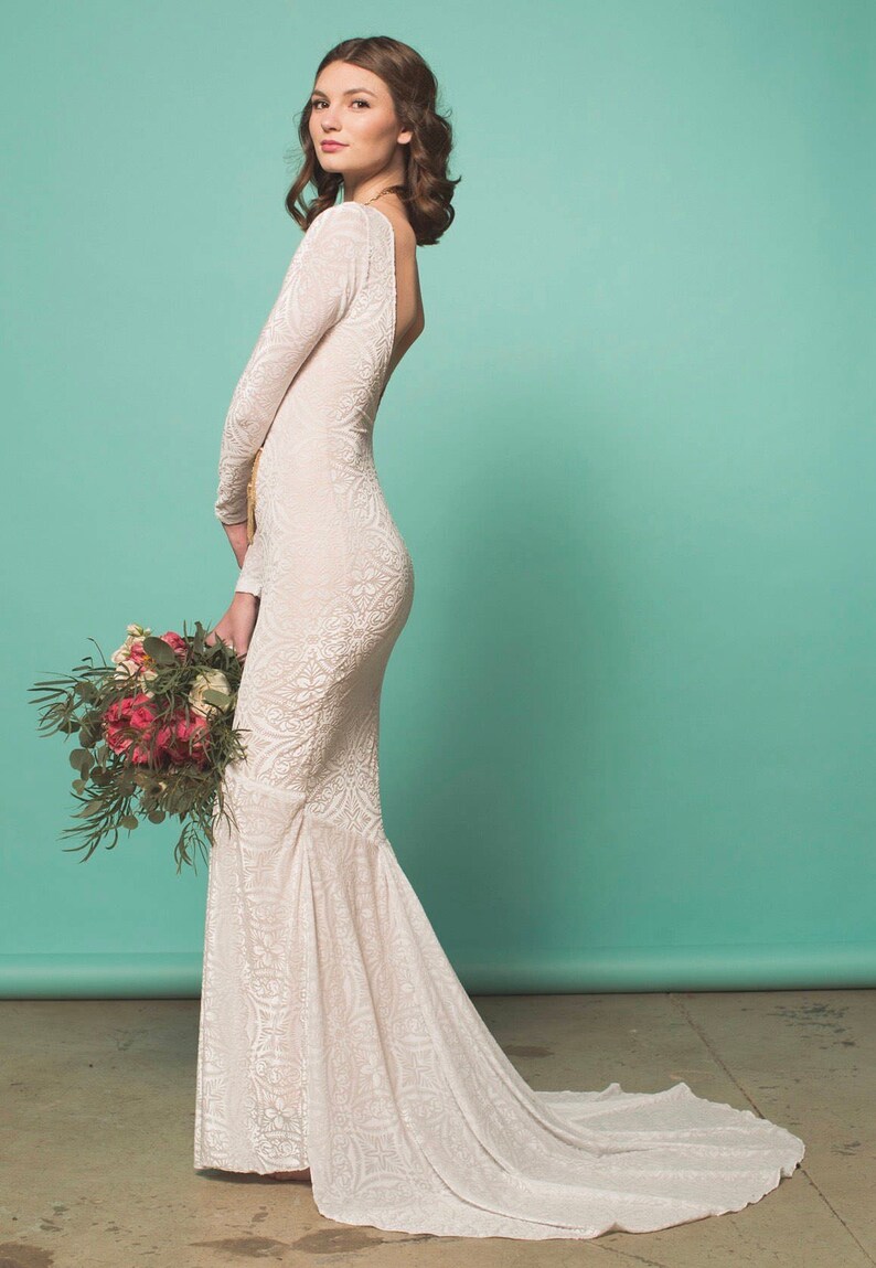May include: A white lace wedding dress with a long sleeve and a low back. The dress has a fitted bodice and a flowing skirt.