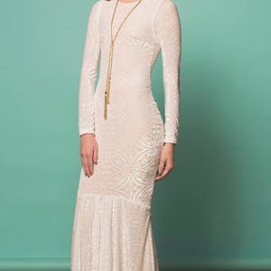 May include: A white long-sleeved maxi dress with a floral crown and a gold chain necklace. The dress has a fitted bodice and a flowing skirt.