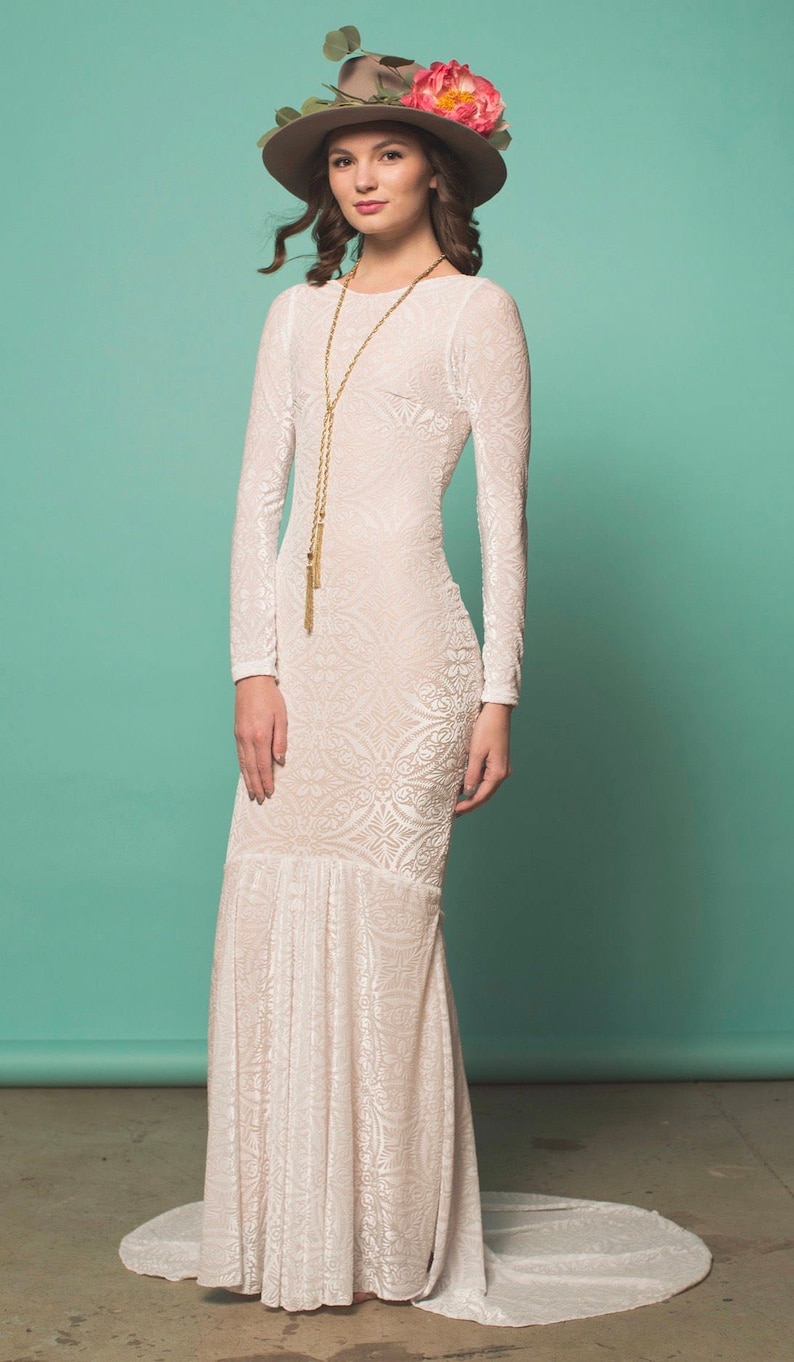 May include: A white long-sleeved maxi dress with a floral crown and a gold chain necklace. The dress has a fitted bodice and a flowing skirt.