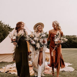 May include: Three women in a field, wearing long dresses and holding bouquets of flowers. The woman in the middle is wearing a white dress and a hat, while the other two women are wearing brown and green dresses. They are walking towards the camera, smiling.