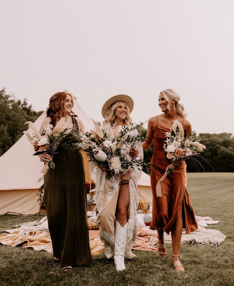 May include: Three women in a field, wearing long dresses and holding bouquets of flowers. The woman in the middle is wearing a white dress and a hat, while the other two women are wearing brown and green dresses. They are walking towards the camera, smiling.