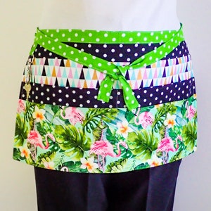 以下が含まれることがあります： 熱帯をテーマにしたカラフルなウエストエプロン。緑色の水玉模様のウエストバンドとリボン、ネイビーブルーの水玉模様、幾何学的な三角形、フラミンゴと花のプリントが特徴です。鍵が側面に付いています。