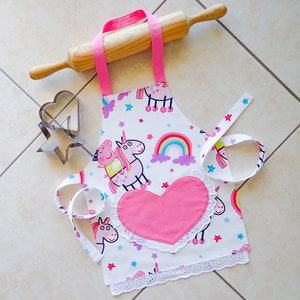 May include: A pink and white children's apron with a heart-shaped pocket and a unicorn and pig design. The apron has a white lace trim and a pink ribbon tie.