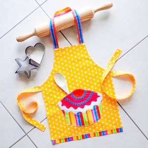Puede incluir: Delantal amarillo de lunares para niños con un diseño de cupcake colorido y una cuchara blanca. El delantal tiene una cinta larga y una cinta más corta.