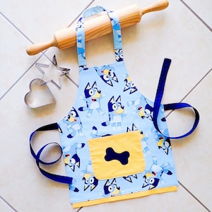 May include: Blue and yellow children's apron with a cartoon dog design. The apron has a large pocket on the front with a bone shape.