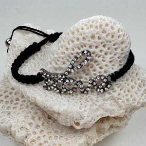 May include: A black cord bracelet featuring a central metal charm in the shape of the word love, encrusted with clear sparkling rhinestones. The bracelet has a braided black cord band with a sliding knot closure and a small silver-toned bead.