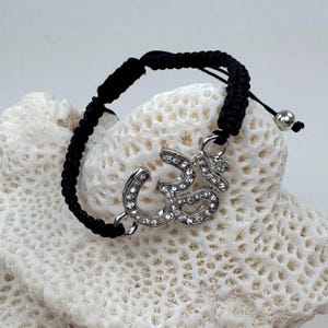 May include: A black cord bracelet featuring a silver-toned Om symbol charm encrusted with clear rhinestones. The bracelet has an adjustable woven cord design with a small silver-toned metal bead at the end of the pull strings.