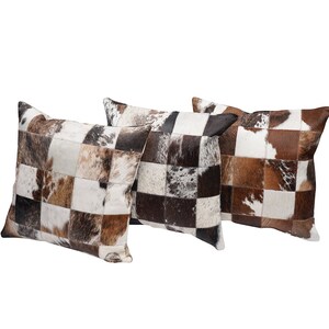 May include: Three square cushions with a patchwork design. The cushions feature a mix of brown, white, and black cowhide squares. The cushions are arranged on a white background.