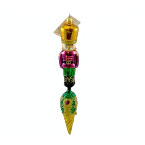 May include: A Christmas ornament shaped like a toy soldier. The ornament has a gold hat, a pink jacket, green pants, and a decorative gold and green base. The ornament is hanging from a tag.