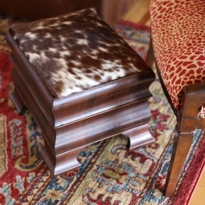 Op de afbeelding: Een rechthoekige houten poef met een koeienhuid bovenkant. De koeienhuid is bruin en wit. De poef heeft een donkerbruin houten frame met decoratieve gesneden details. De achtergrond omvat een patroonkleed en een stoel.