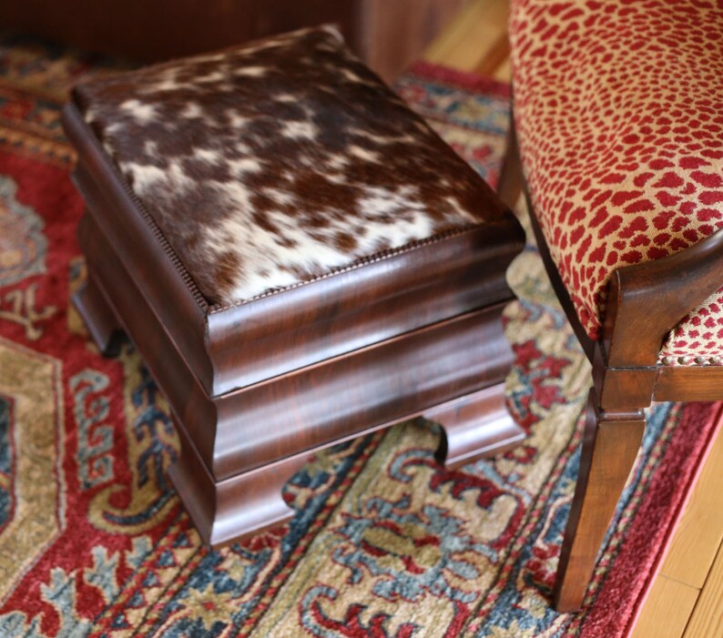 Op de afbeelding: Een rechthoekige houten poef met een koeienhuid bovenkant. De koeienhuid is bruin en wit. De poef heeft een donkerbruin houten frame met decoratieve gesneden details. De achtergrond omvat een patroonkleed en een stoel.
