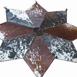 May include: A brown, black, and white cowhide rug in the shape of a star with a brown leather center. The rug has a fringe around the edges.