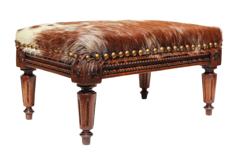 Puede incluir: Un otomano de madera marr&oacute;n con tapa de piel de vaca y ribete de tachuelas doradas. El otomano tiene cuatro patas torneadas.