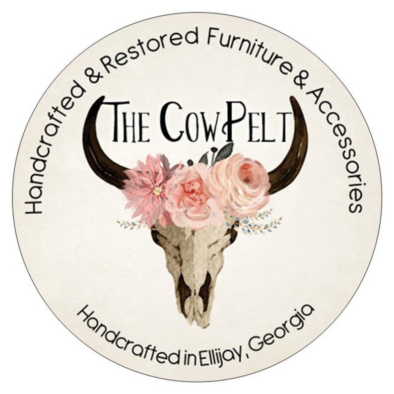 May include: A white circle with a black cow skull with a floral crown of pink and white flowers. The text "The Cow Pelt" is above the skull. The text "Handcrafted & Restored Furniture & Accessories" is around the top of the circle. The text "Handcrafted in Ellijay, Georgia" is around the bottom of the circle.