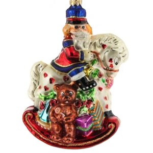 May include: A Christmas ornament featuring a nutcracker soldier riding a white rocking horse decorated with red hearts and green foliage. A brown teddy bear and a purple gift box are at the base of the horse. The ornament has a red and gold base.