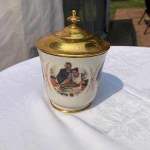 May include: A white ceramic jar with a gold lid and a painted image of a family gathered around a table. The image depicts a man, woman, and two children. The jar has a gold trim around the base.
