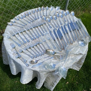 May include: A set of silver flatware, including forks, knives, spoons, and serving pieces, arranged on a white tablecloth. The flatware is laid out in rows, with the handles facing up. The flatware is in a plastic bag.