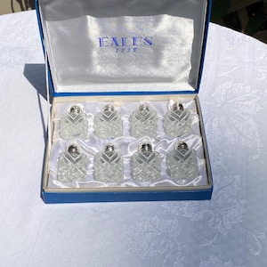 May include: A set of seven clear glass salt shakers with silver tops in a blue box. The box has the text "Eailes 1779" on the inside lid.