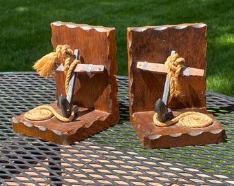 May include: A pair of wooden bookends with a nautical theme. Each bookend features a carved wooden anchor with a rope wrapped around it. The anchors are painted black and the rope is a light brown color.