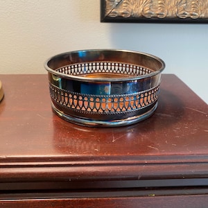 May include: Silver-plated wine bottle holder with an intricate design. The holder has a round shape and a decorative border.