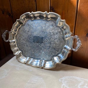 May include: Silver-plated serving tray with ornate floral detailing. The tray has a scalloped edge and two handles. The interior features a rectangular area for engraving. The tray is displayed on a cream-colored surface with a bunny pattern, against a wooden background.