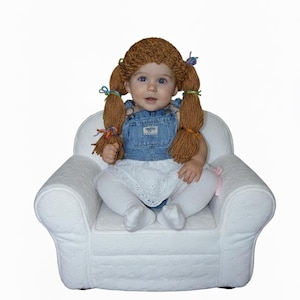 Op de afbeelding: Een baby verkleed als pop, compleet met een bruine pruik van garen met vlechten vastgebonden met kleurrijke linten. De baby draagt een spijkerbroek, een witte oogjesrok en witte sokken, zittend in een kleine witte gestoffeerde fauteuil.