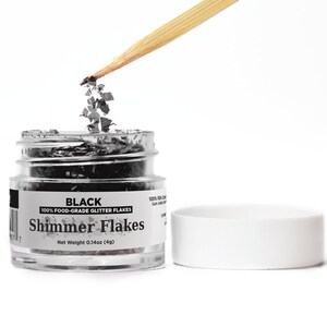 May include: A small clear jar filled with black food-grade glitter flakes. The jar has a white label with the text "BLACK 100% FOOD-GRADE GLITTER FLAKES Shimmer Flakes Net Weight 0.14oz (4g)".