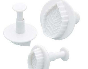 May include: Three white plastic leaf-shaped cookie cutters with a plunger handle. The cutters are designed to create leaf shapes for decorating cakes and cookies.