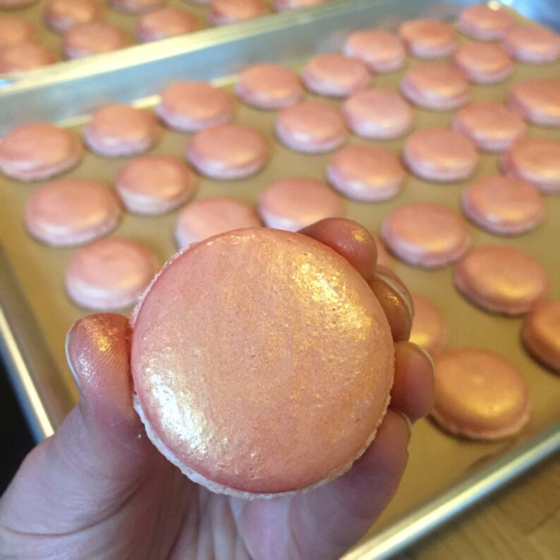 May include: A close-up of a single pink macaron with a shimmery, pearlescent finish. The macaron is on a baking sheet with other macarons in the background.