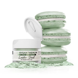 May include: A jar of shimmering, edible Chrysanthemum Luster Dust with a net weight of 0.14oz (4g) next to a stack of four light green macarons with white filling. The jar's lid is open, and a small pile of dust is visible.