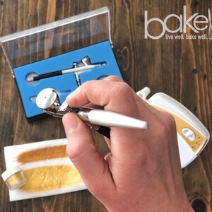 May include: A silver airbrush gun with a gold nozzle is held in a hand. The airbrush is part of a set that includes a small air compressor and a container of gold dust. The airbrush set is labeled "bakell live well. bake well."