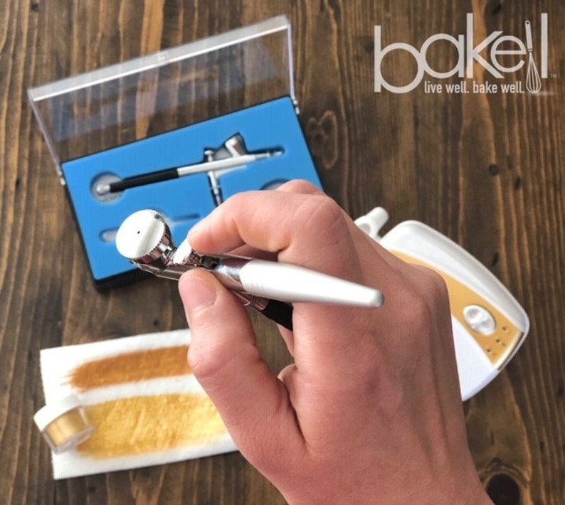 May include: A silver airbrush gun with a gold nozzle is held in a hand. The airbrush is part of a set that includes a small air compressor and a container of gold dust. The airbrush set is labeled "bakell live well. bake well."