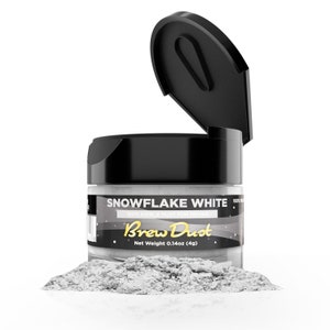 May include: A small jar of white edible dust called "Snowflake White" by Brew Dust. The jar is open and the dust is spilling out onto a white surface. The jar label says "100% Edible Dust for Drinks" and "Net Weight 0.14oz (4g)".