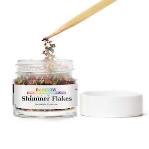 May include: A small glass jar filled with colorful iridescent glitter flakes. The jar has a white label with the text "RAINBOW 100% FOOD-GRADE GLITTER FLAKES Shimmer Flakes Net Weight 0.14oz (4g)".