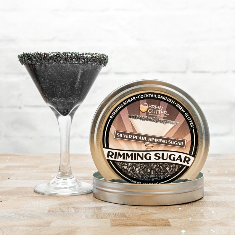 May include: A silver pearl rimming sugar in a round tin container with the text "Brew Glitter" and "Silver Pearl Rimming Sugar" on the label. The container is sitting on a wooden surface next to a martini glass with a black rim.