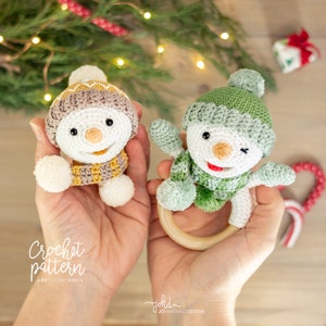 May include: Two crocheted snowman ornaments, one in brown and beige with a yellow scarf and the other in green with a green scarf. Both have white faces with black eyes and a black nose. The green snowman is attached to a wooden ring.