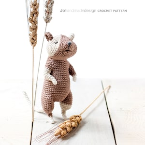 Pode incluir: Um brinquedo de rato de crochê marrom e branco em pé sobre uma superfície de madeira branca com dois talos de trigo.
