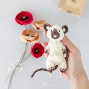 Puede incluir: Juguete de amigurumi de ratón tejido a crochet con un cuerpo blanco, orejas marrones y una nariz marrón. El juguete se sostiene en una mano sobre un fondo blanco.