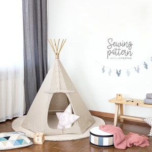 Könnte beinhalten: Ein beigefarbenes Tipi-Zelt mit einem Holzpfahl an der Spitze. Das Tipi steht in einem Raum mit Holzboden und einer weißen Wand. An der Wand hängt eine Kaktusgirlande. Der Text "Sewing pattern MEDIUM TEEPEE" ist an der Wand geschrieben.
