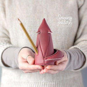 May include: A red fox-shaped pen holder made of leather with a pencil sticking out of the top. The text "Sewing pattern FOXPENHOLDER" is visible above the fox.