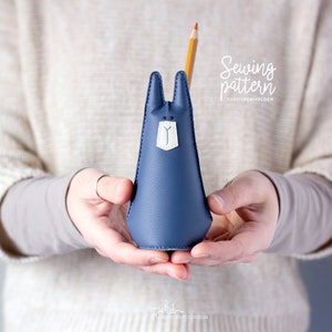 Pode incluir: Um porta-canetas de coelho de feltro azul com detalhes em feltro branco. O coelho está segurando um lápis na boca. O texto "Sewing pattern" e "Rabbit Penholder" é visível na imagem.