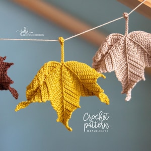 Könnte beinhalten: Gehäkelte Ahornblattdekorationen in Gelb-, Braun- und Beigetönen hängen an einer Schnur. Die Blätter sind mit sichtbaren Nähten versehen und vor einem sanften blauen Hintergrund aufgehängt. Der Text "Crochet pattern MAPLELEAVES" ist sichtbar.