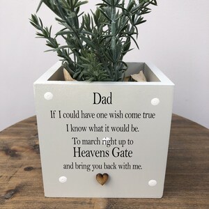 May include: A square, light gray planter with the word "Dad" and a heartfelt message. The planter is adorned with a small wooden heart and white decorative accents. It holds a green plant, creating a touching memorial.